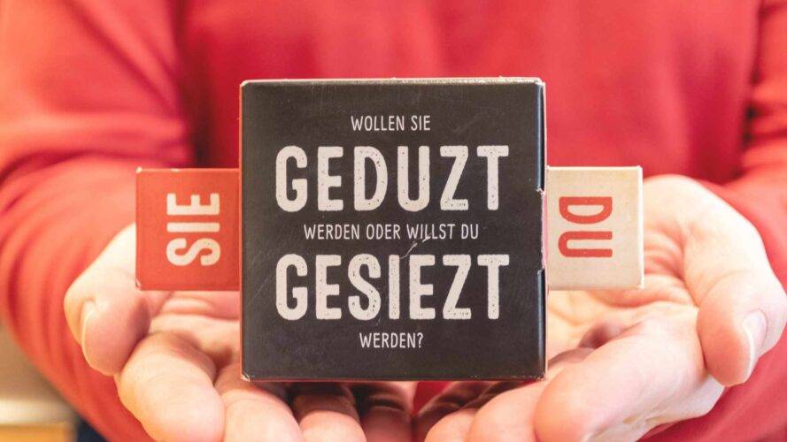 Schwarzer Würfel mit „Sie“ und „Du“-Optionen in Händen.